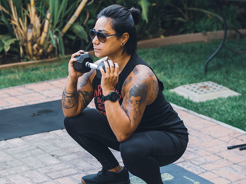 Person wearing sunglasses performing a squat exercise outdoors with a dumbbell in each hand, showing tattooed arms and athletic wear.